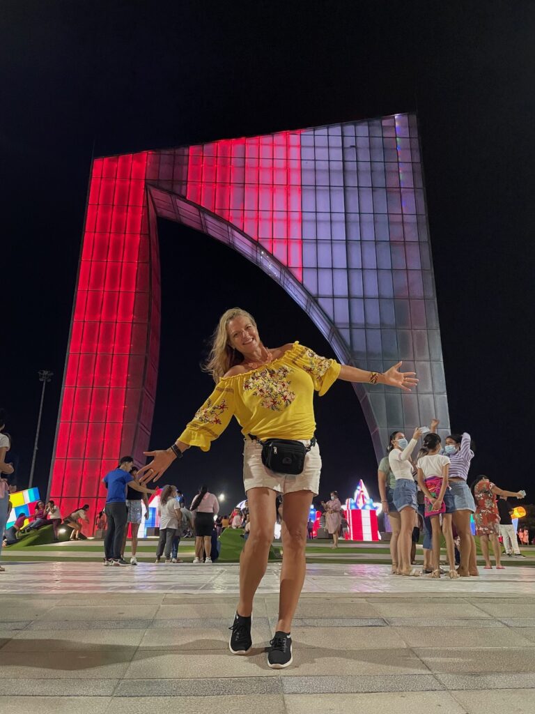 Travel blogger Kate Dana stands smiling with open arms during a nighttime visit to the Aleta de Juniors monument in Barranquilla, Colombia photo ©Kate Dana